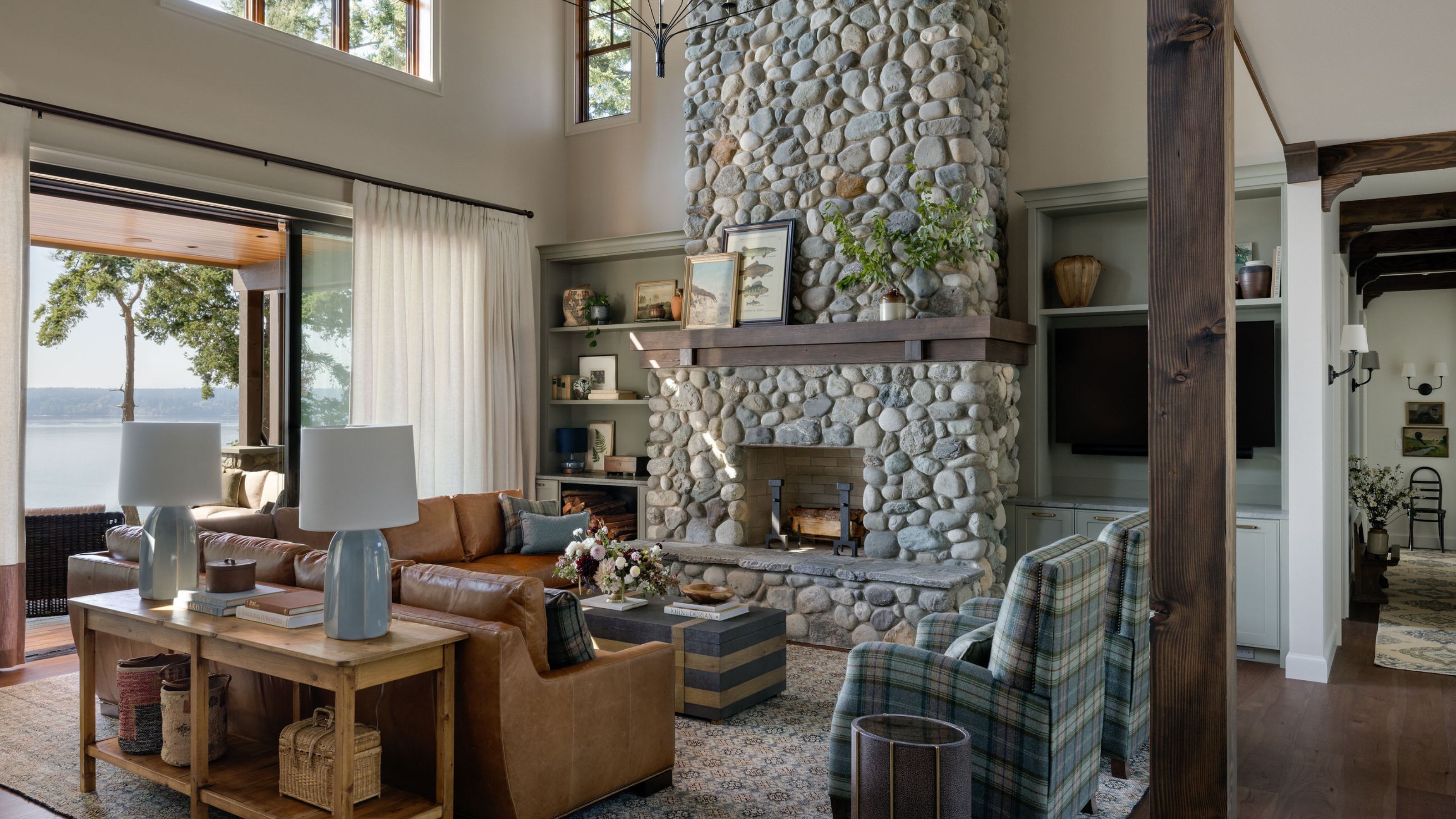 A living room with tall sloped ceilings, a statement stone fireplace, warm white walls, sliding glass doors, a leather sectional sofa and a wooden console table, and glass sliding doors. 
