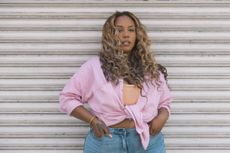 A woman with long curly hair poses against a white metal roller shutter. Dressed in a pastel pink button-up shirt and light denim jeans, she exudes confidence and casual elegance in an urban setting.