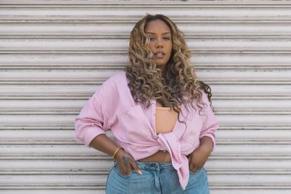 A woman with long curly hair poses against a white metal roller shutter. Dressed in a pastel pink button-up shirt and light denim jeans, she exudes confidence and casual elegance in an urban setting.