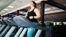 woman walking on a treadmill with earphones in, she's in a gym setting wearing turquoise leggings and black vest.