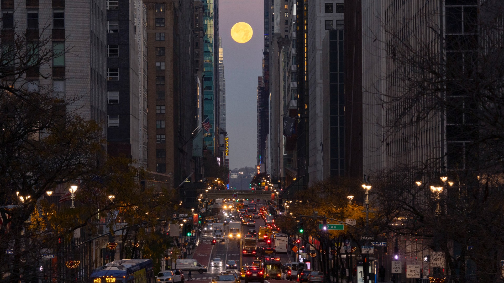 La luna llena se puede ver sobre una calle muy transitada entre dos filas de grandes edificios en una ciudad abarrotada de gente.