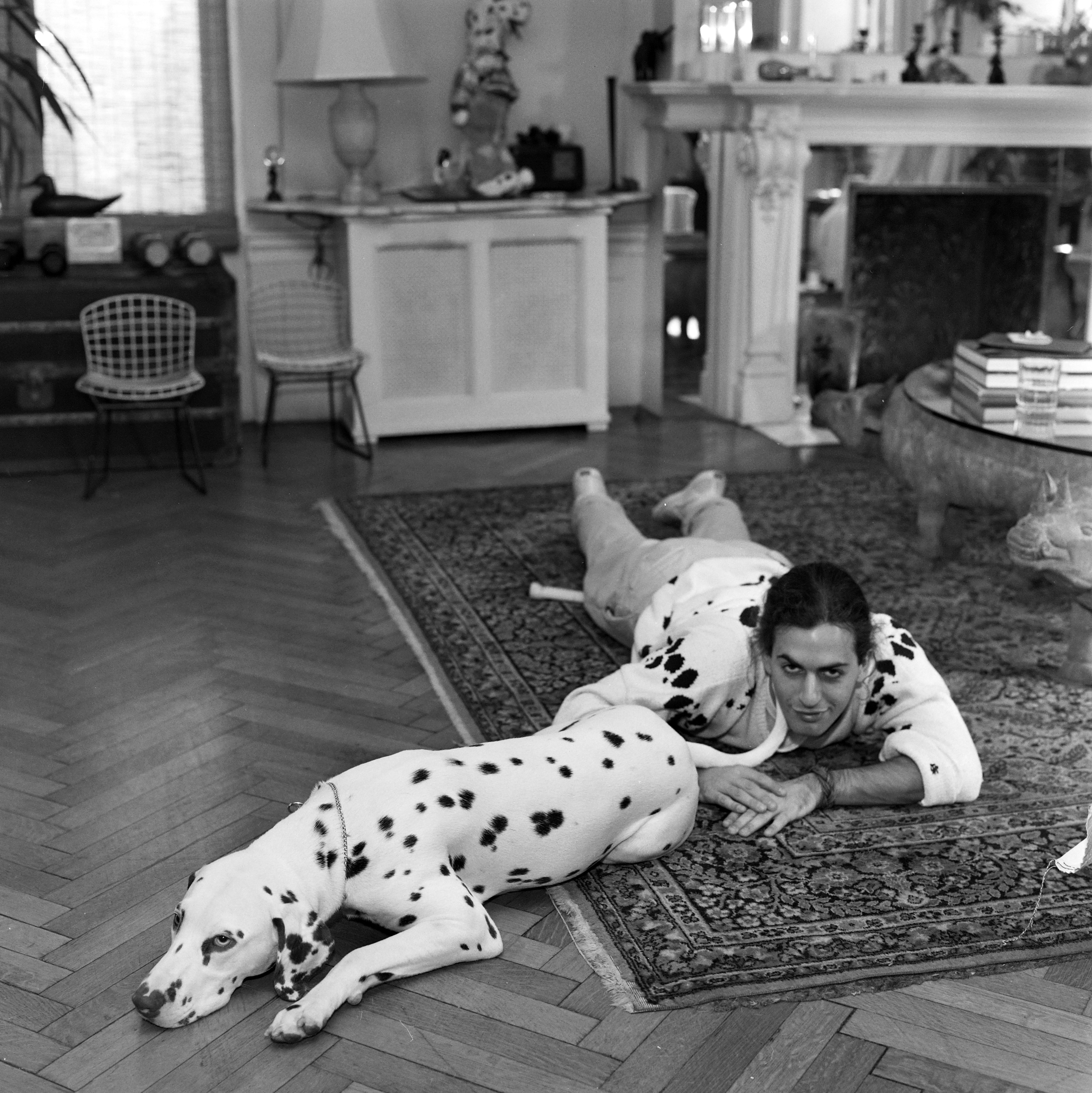 A black-and-white photograph of Marc Jacobs lying on a patterned rug beside a resting dalmatian in a modern living room with a fireplace.