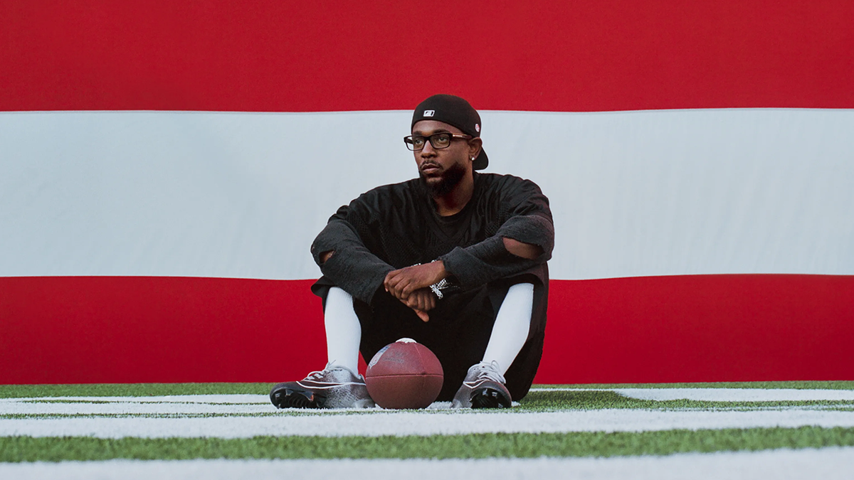 Kendrick Lamar sits on a football field with a ball, promoting his Super Bowl Halftime Show