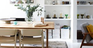 a white open-plan living room diner with a wooden table and alcove shelves filled with decorative glass items