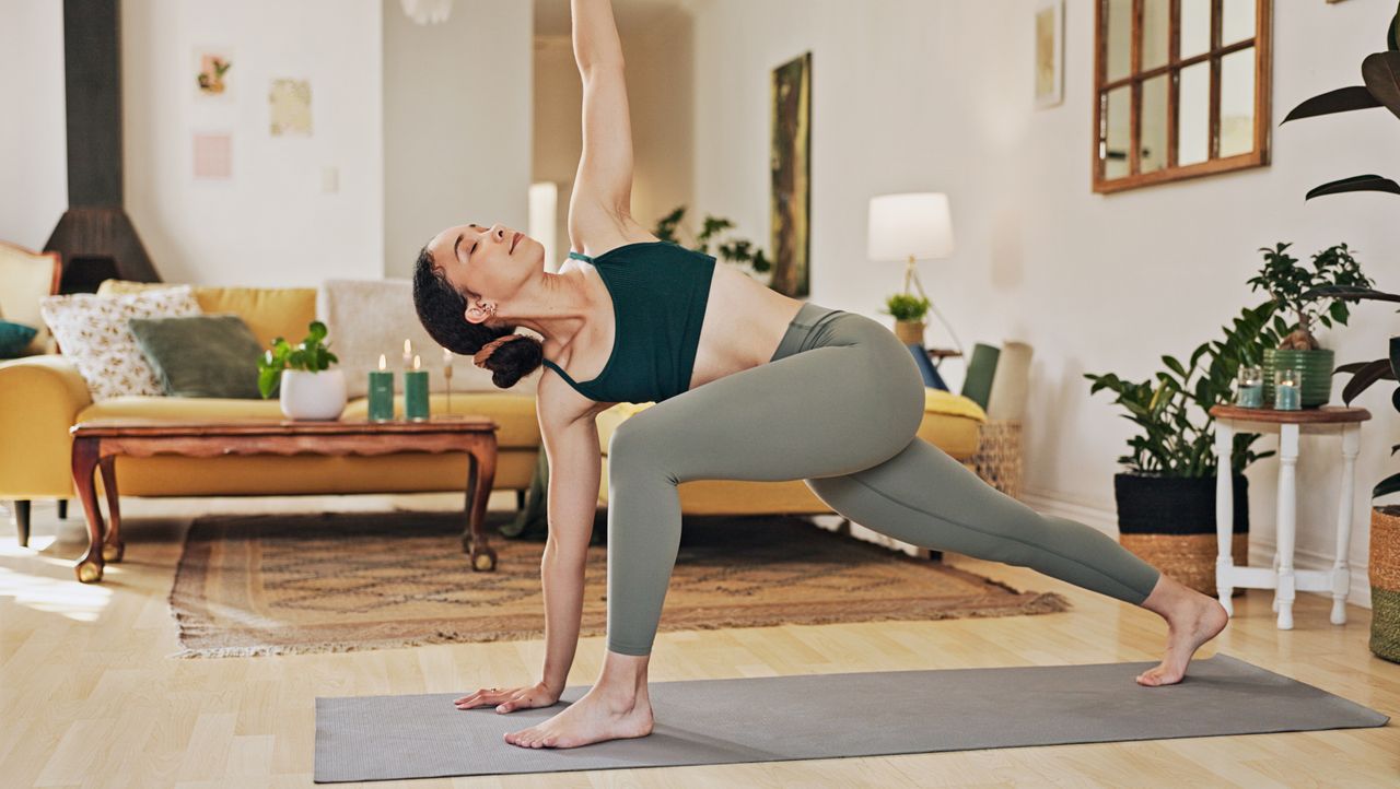 Woman stretching at home. One foot and the opposite hand are in line on the floor at the top of a yoga mat, with the other foot on its toes at the back of the mat. The other arm reaches up and her torso is rotated. She is wearing green sports leggings and a cropped black top and her black hair is in a bun.