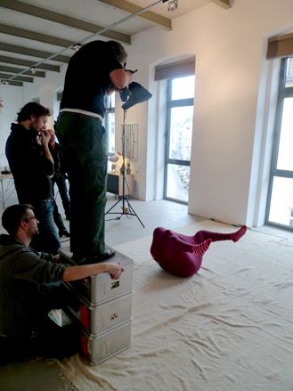 Photographer standing on a pile of boxes to take a photo of a someone on their back in a bodysuit with no opening for the arms or head