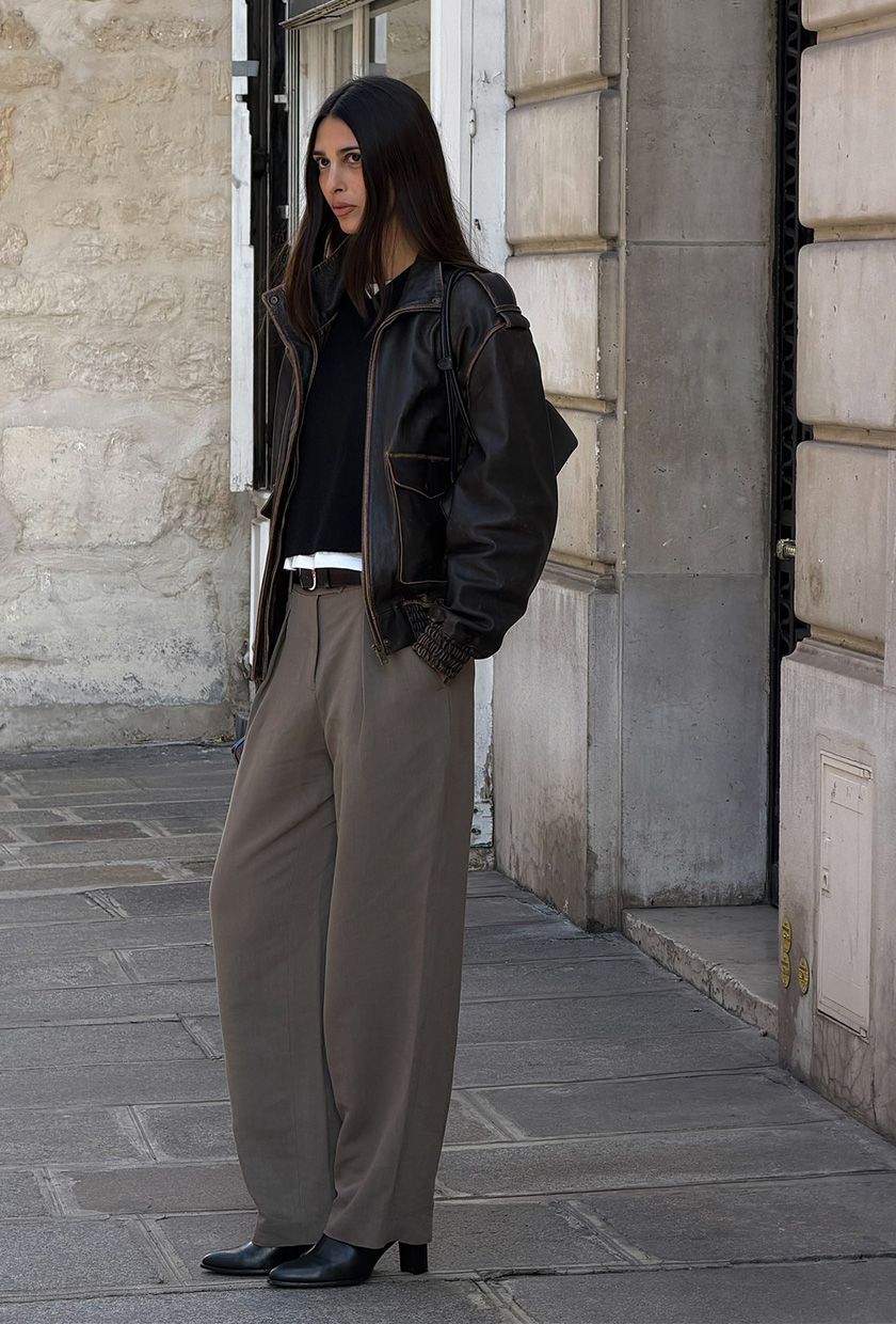 A woman wearing a brown leather bomber jacket over a black sweater and white T-shirt, styled with gray trousers and black ankle boots.