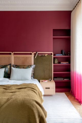 Close-up image of a magenta-pink painted bedroom. The bed has a wooden, rattan, and green headboard. The bedding is white and olive green. There are built-in shelves on the wall behind the bed and the curtain is white and magenta ombre.