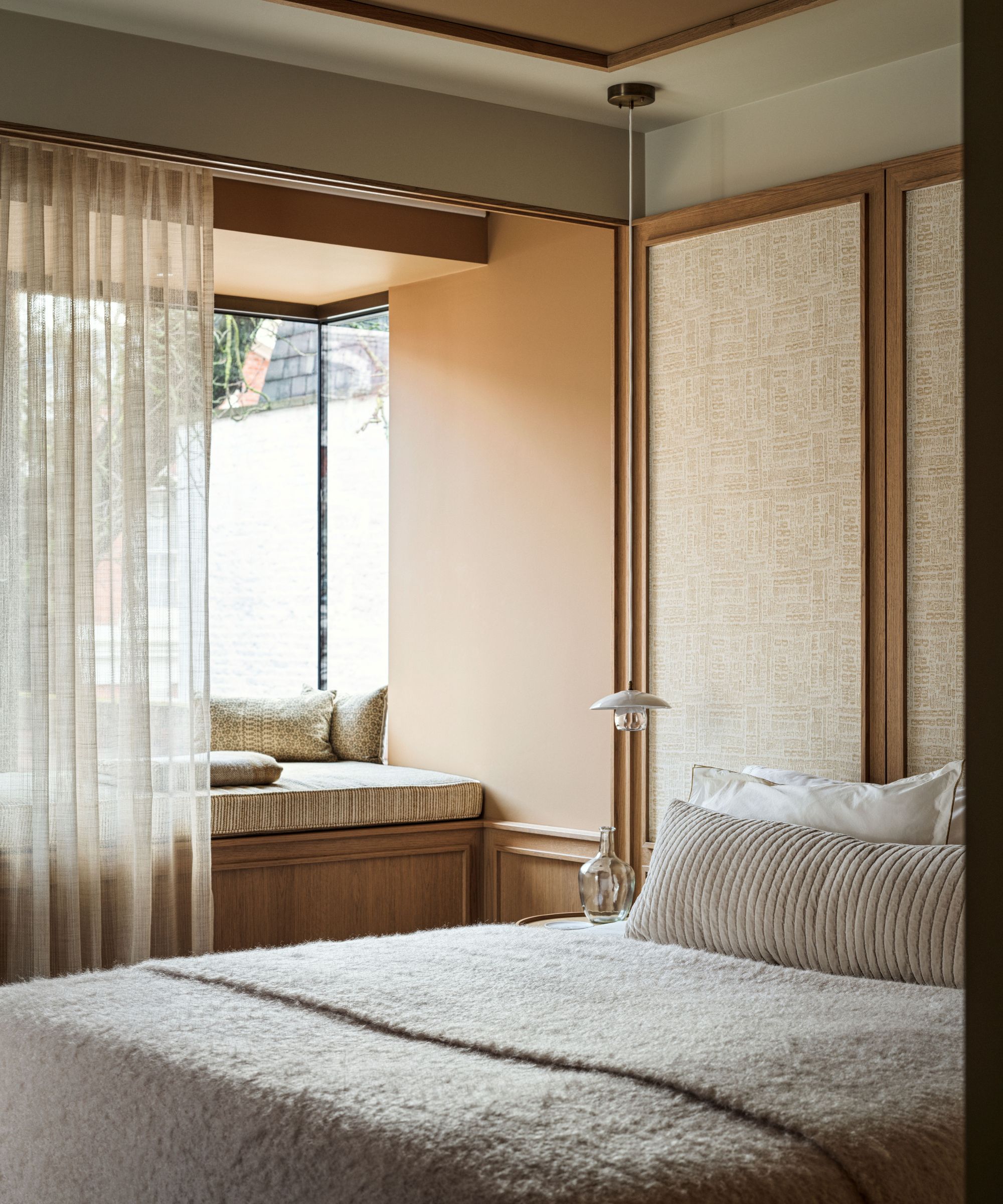 Warm neutral bedroom, a bed with a beige fluffy throw and a striped bolster cushion and a window seat with sheer drapes.