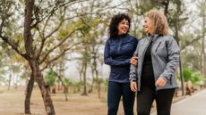Two women in workout tracksuits walking arm in arm in a park smiling at eachother