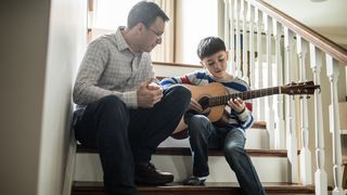 Man watches his son play acoustic guitar while they both sit on a staircase