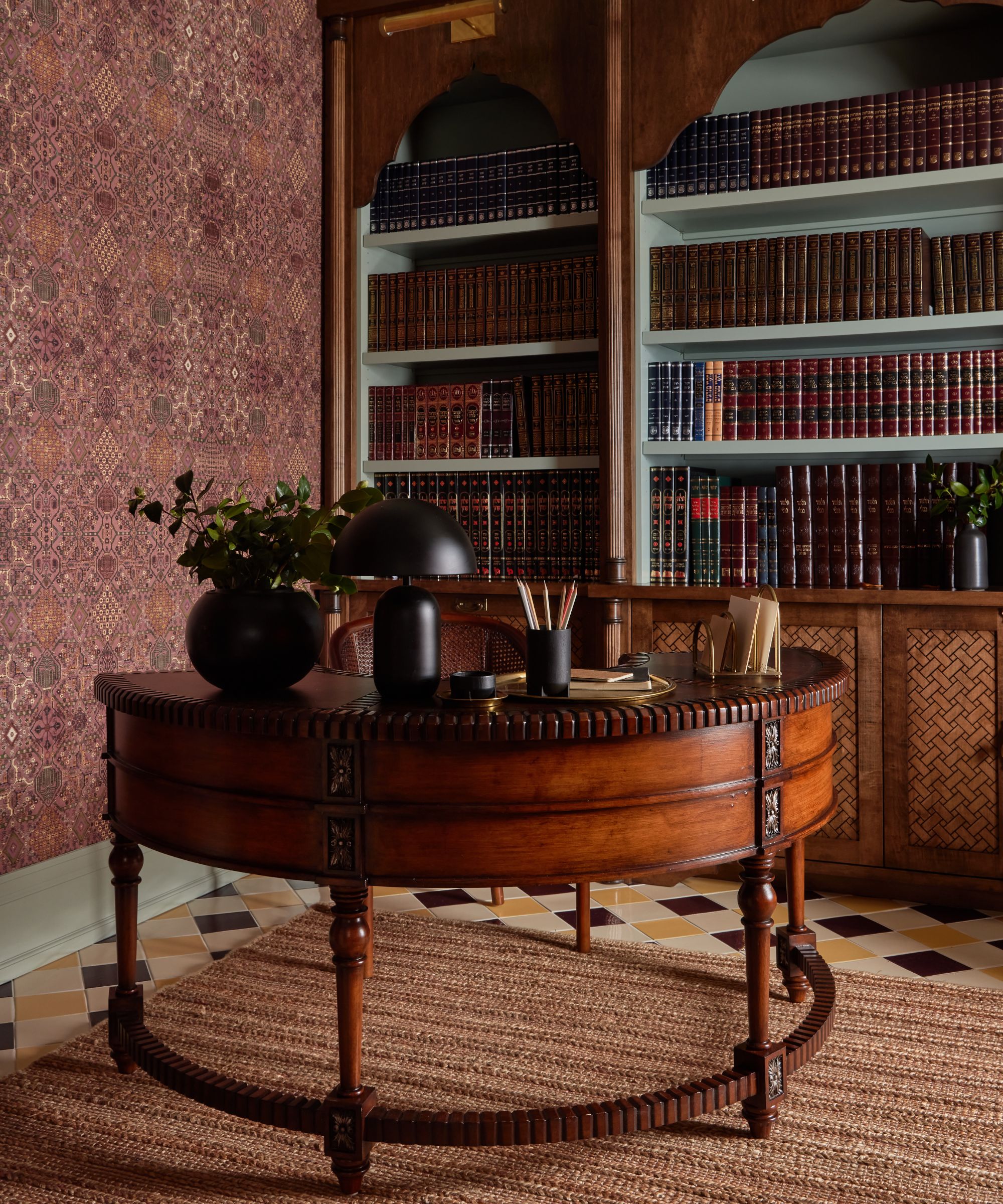 Close up of round, dark wood antique desk in study, decorated with lamp, ornaments and papers, shelves with books in background, patterned wallpaper, tiled floor, rug under table