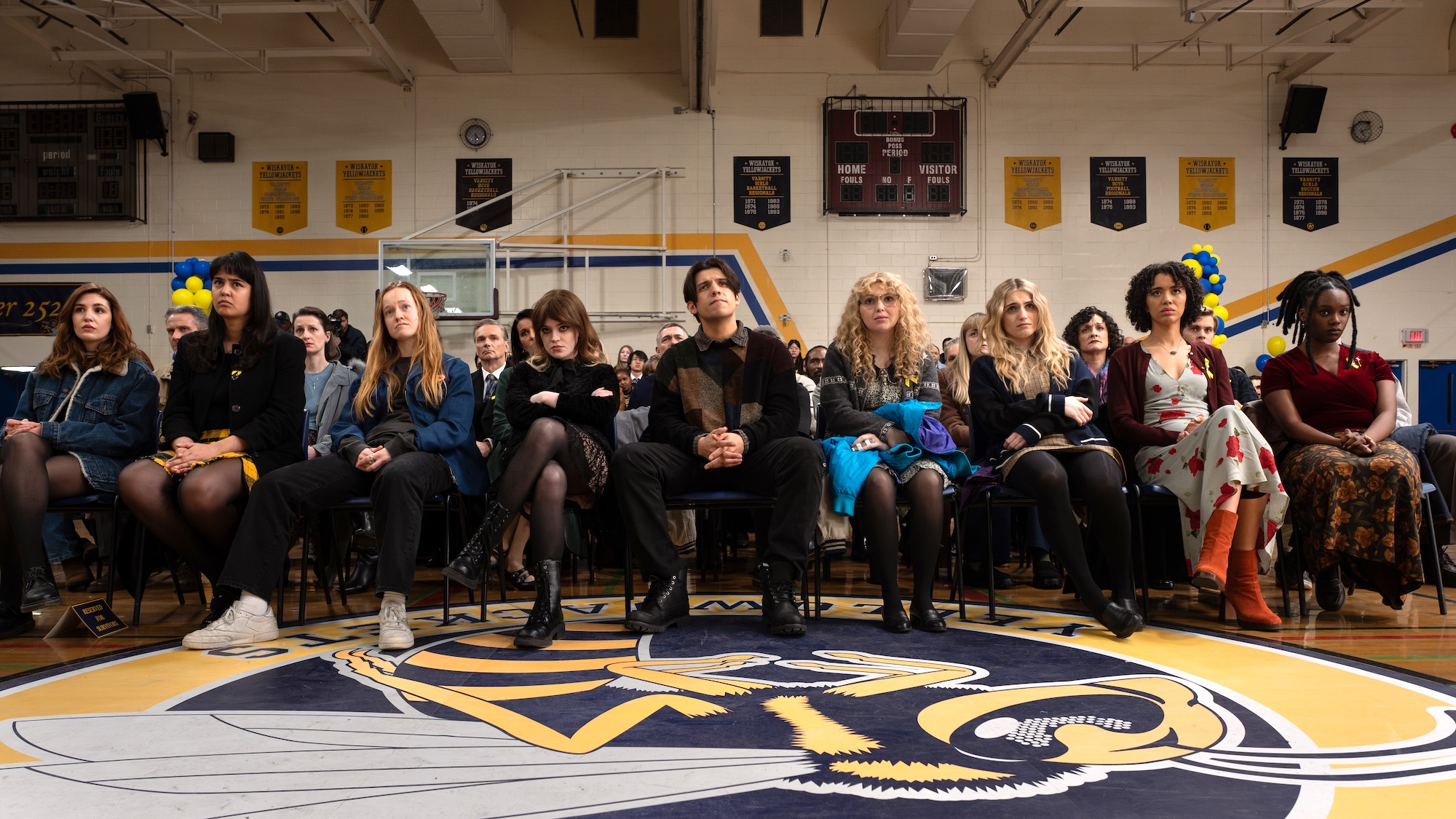 Shauna, Lottie, Van, Natalie, Travis, Misty, Melissa, Taissa, and Akilah sitting in a line of chairs in the middle of their high school gym after being rescued in Yellowjackets Season 4