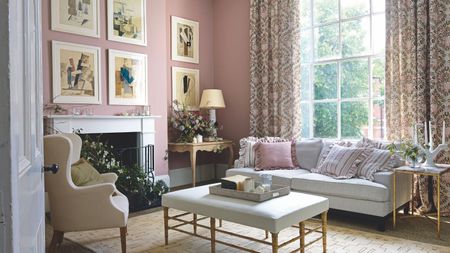 A vintage style pink living room with patterned curtains in front of a large window, with white framed gallery wall, a white mantelpiece, a grey sofa with frilly pink and striped cushions with matching footrest with tray and a blush pink armchair facing towards it