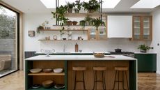 Lots of greens and woody textures have been used in this kitchen. Hanging about the island are a brunch of plants stacked on shelves and below and wooden bar stools with island storage to the left.