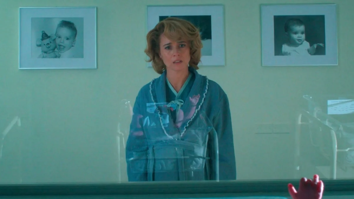 Linda Cardellini standing in front of a crib behind a glass window, looking shocked