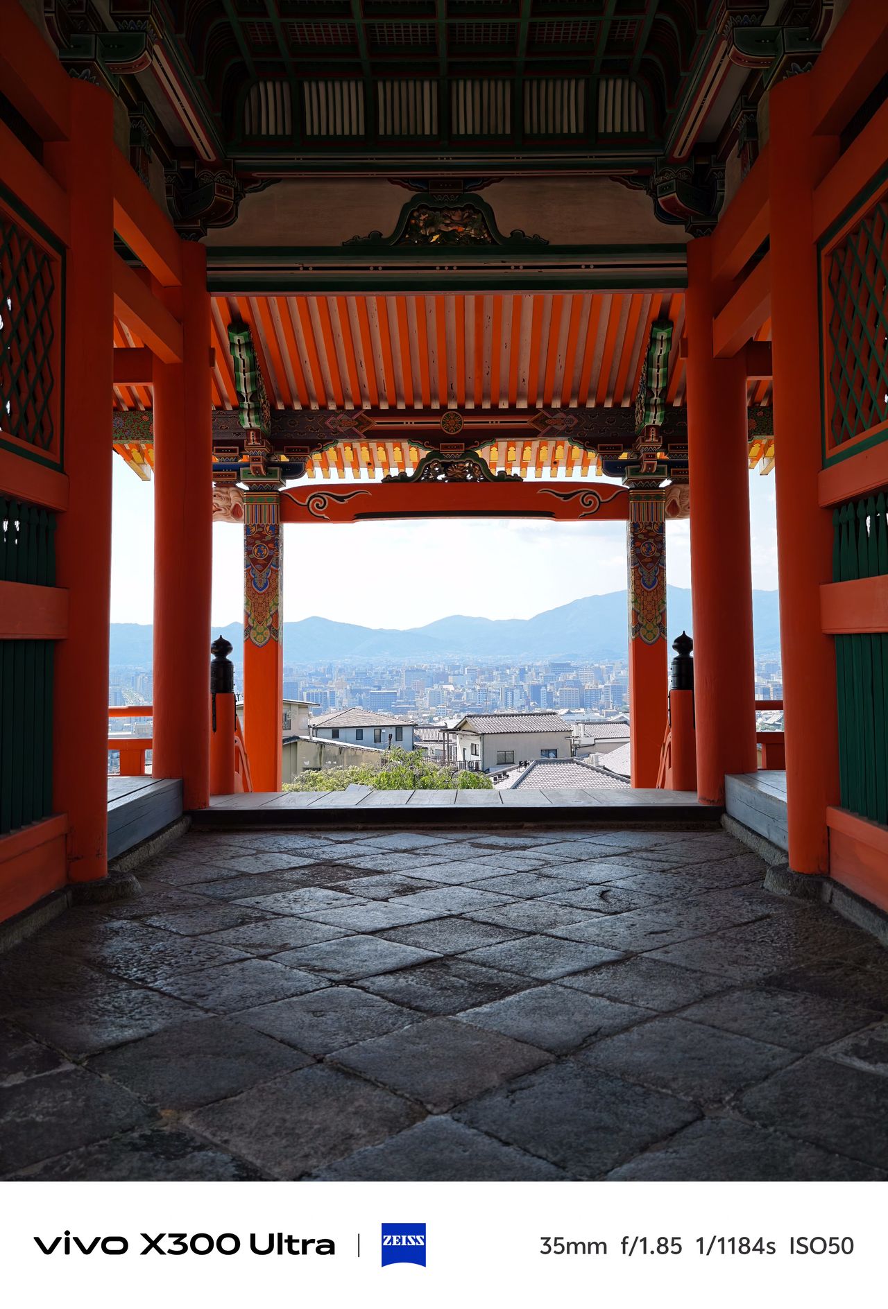 View through an ornate orange wooden temple gate looking out onto a dense cityscape stretching toward distant blue mountains.
