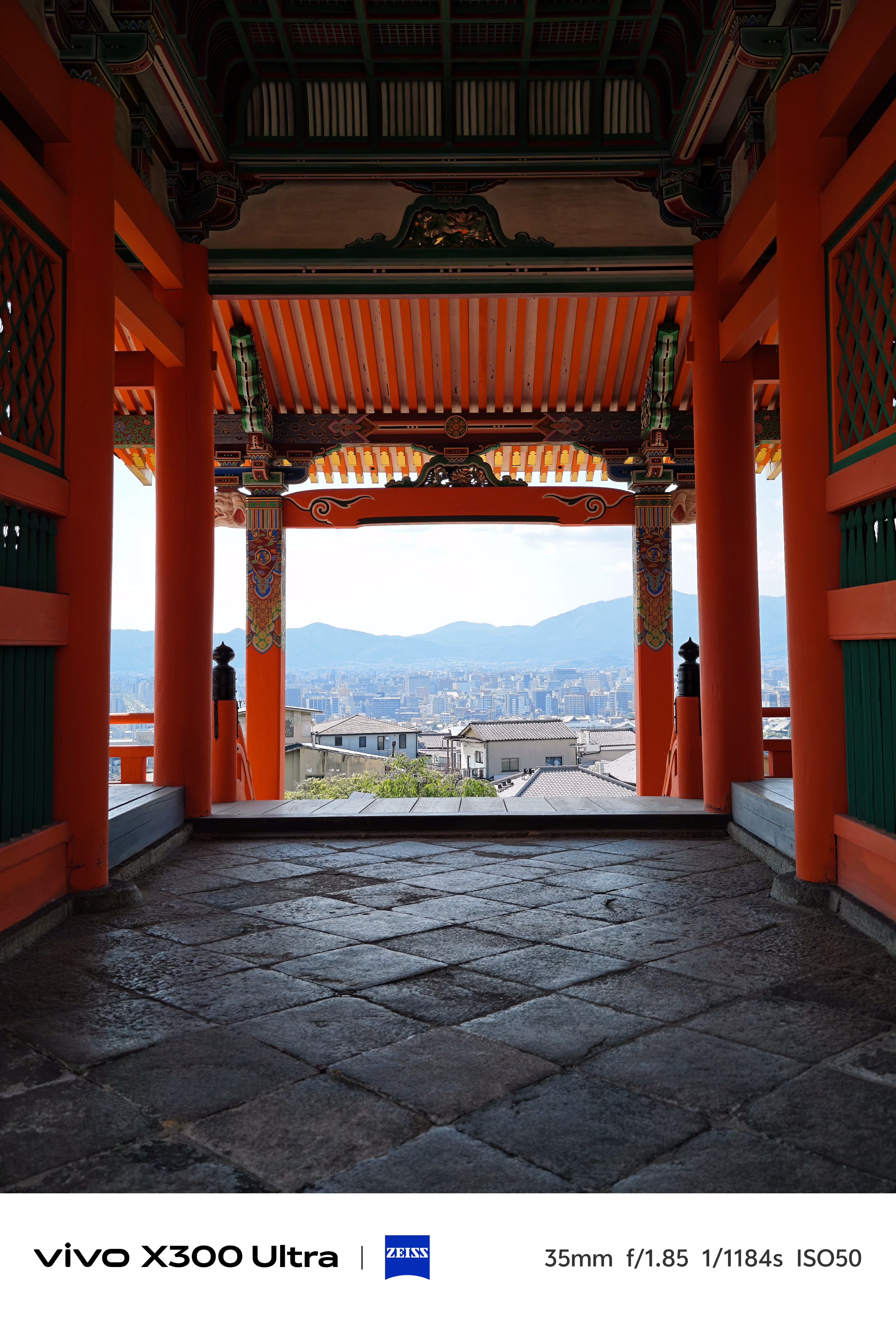 View through an ornate orange wooden temple gate looking out onto a dense cityscape stretching toward distant blue mountains.