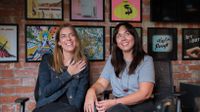 two women smiling with a brick wall covered in artwork behind them