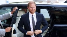 Prince Harry, wearing a navy blue suit and smiling, steps out of a black car
