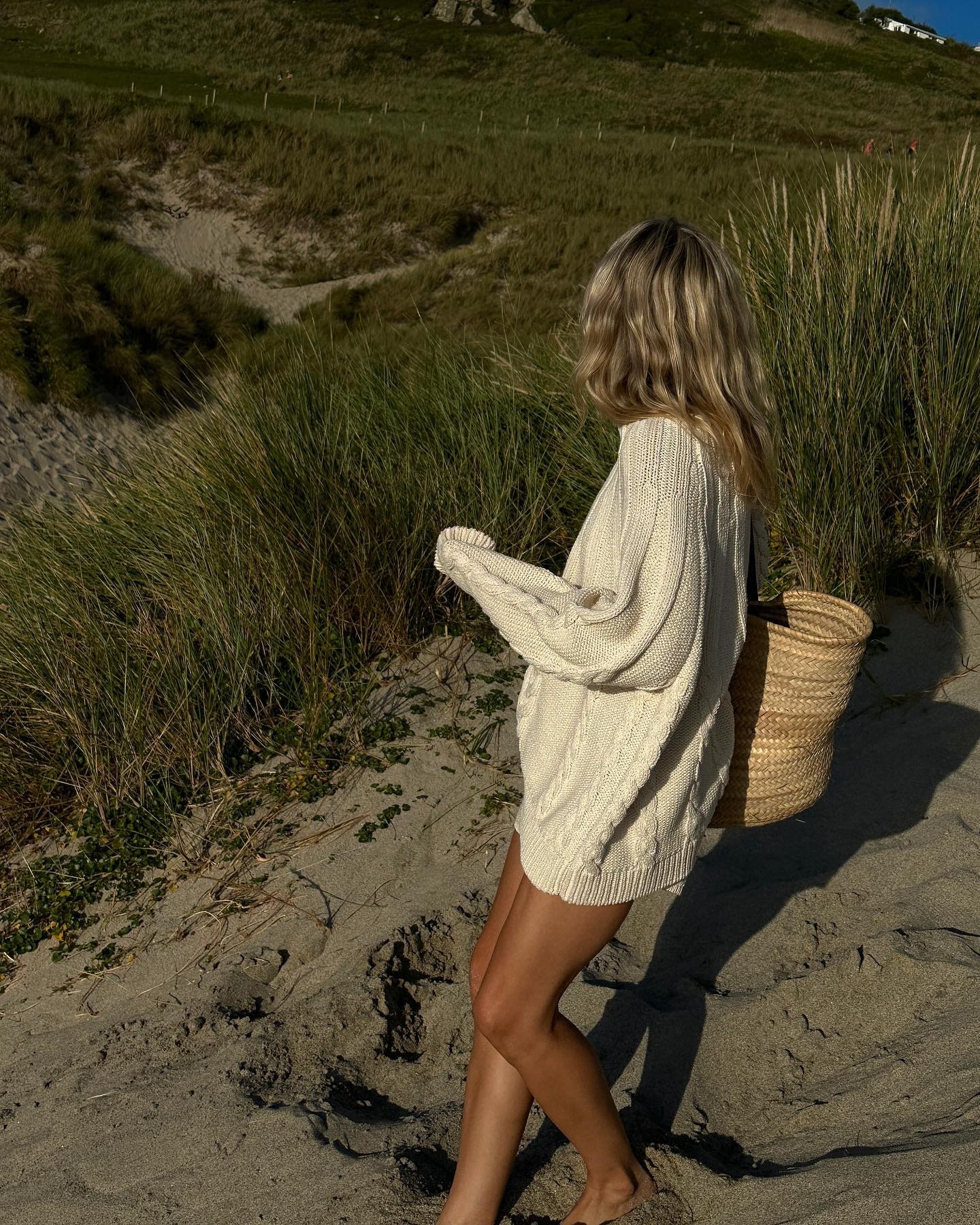 Lucy Williams wearing a cotton loose sweater with a straw bag on the beach.