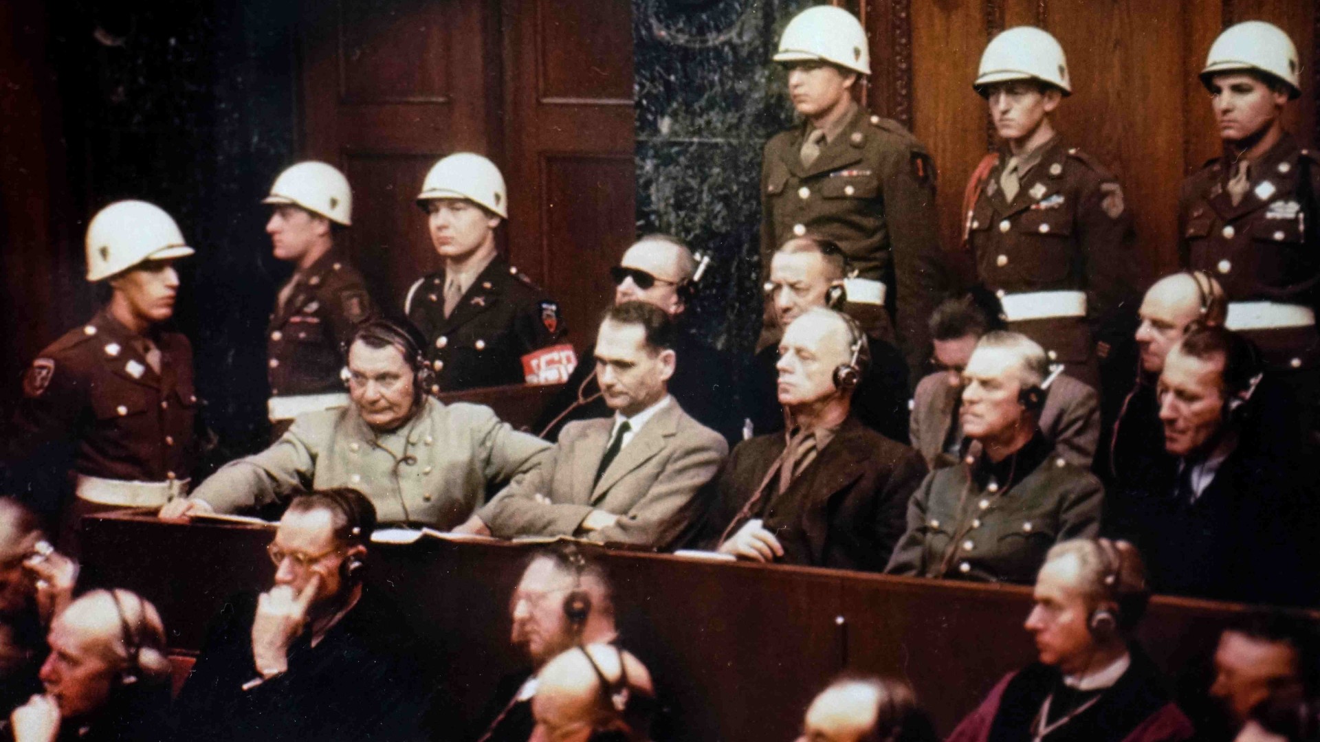 Photograph of Rudolf Hess taken during the Nuremberg Trials. He is sitting in the middle of a bench with his arms crossed, wearing a light tan suit with a black tie. He has short black hair. Behind him are six guards wearing uniform and white helmets. Either side of him and on the bench below are a number of men in suits all wearing headphones.