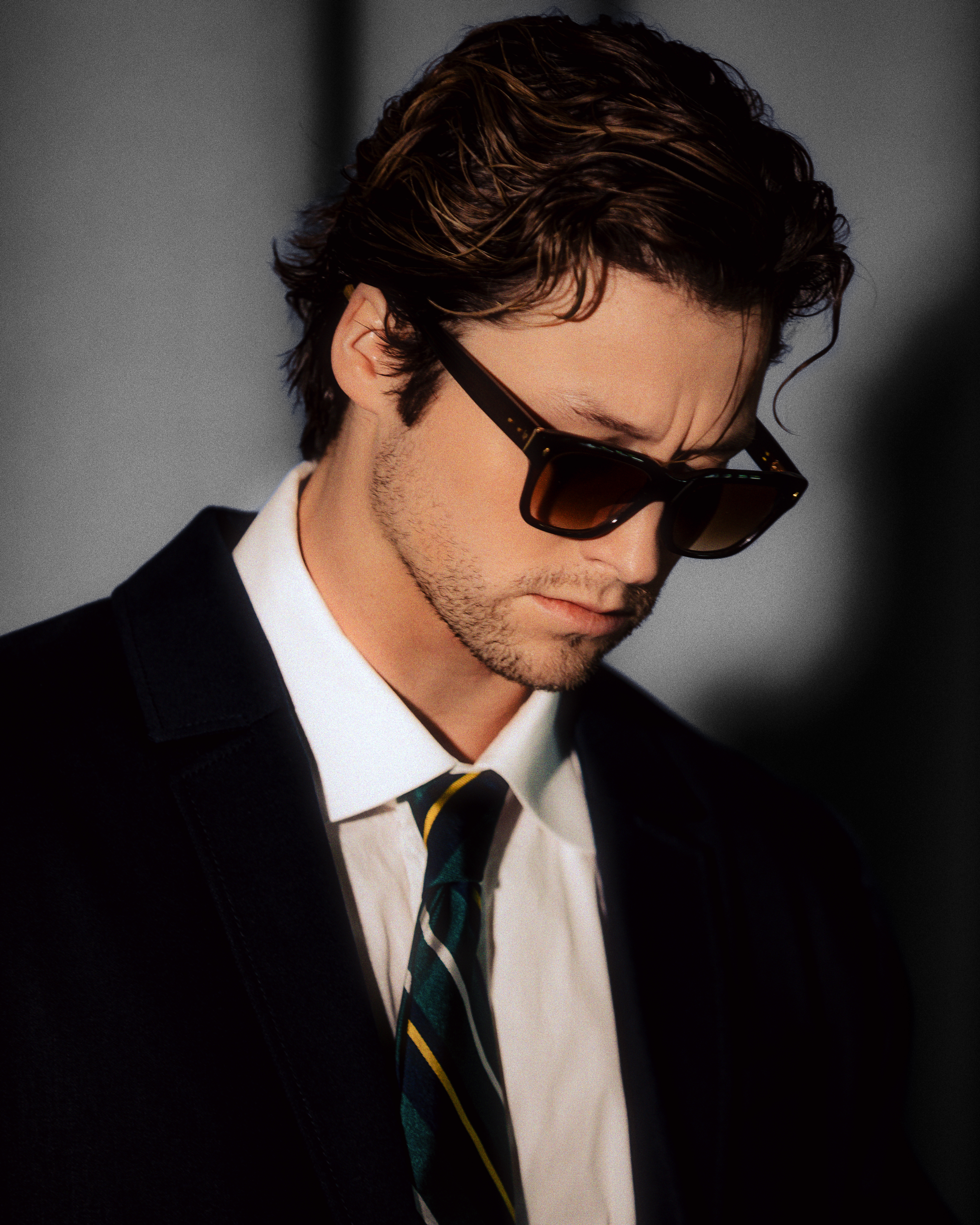 Off Campus star Stephen Kalyn wearing sunglasses, a black jacket, a white button-down shirt, and a striped tie.