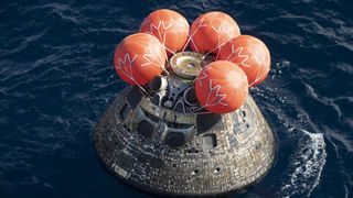a charred cone-shaped spacecraft floats in the ocean with five large red spherical balloons on top