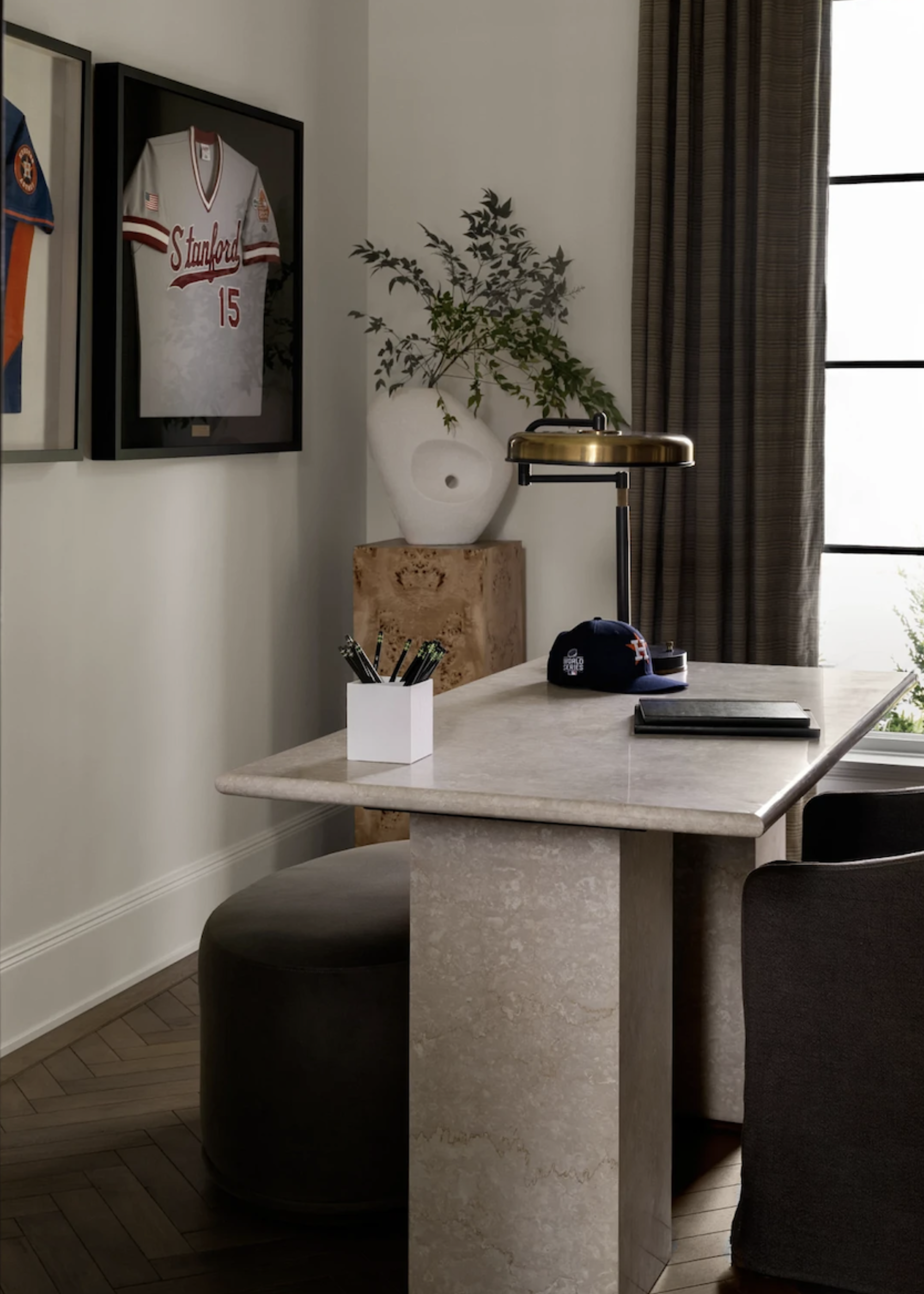 A minimalist home office with a marble desk, a dark armchair, a soft stool, a brass table lamp, a stationery holder, a burl plinth with a vase of stems, and framed shirts