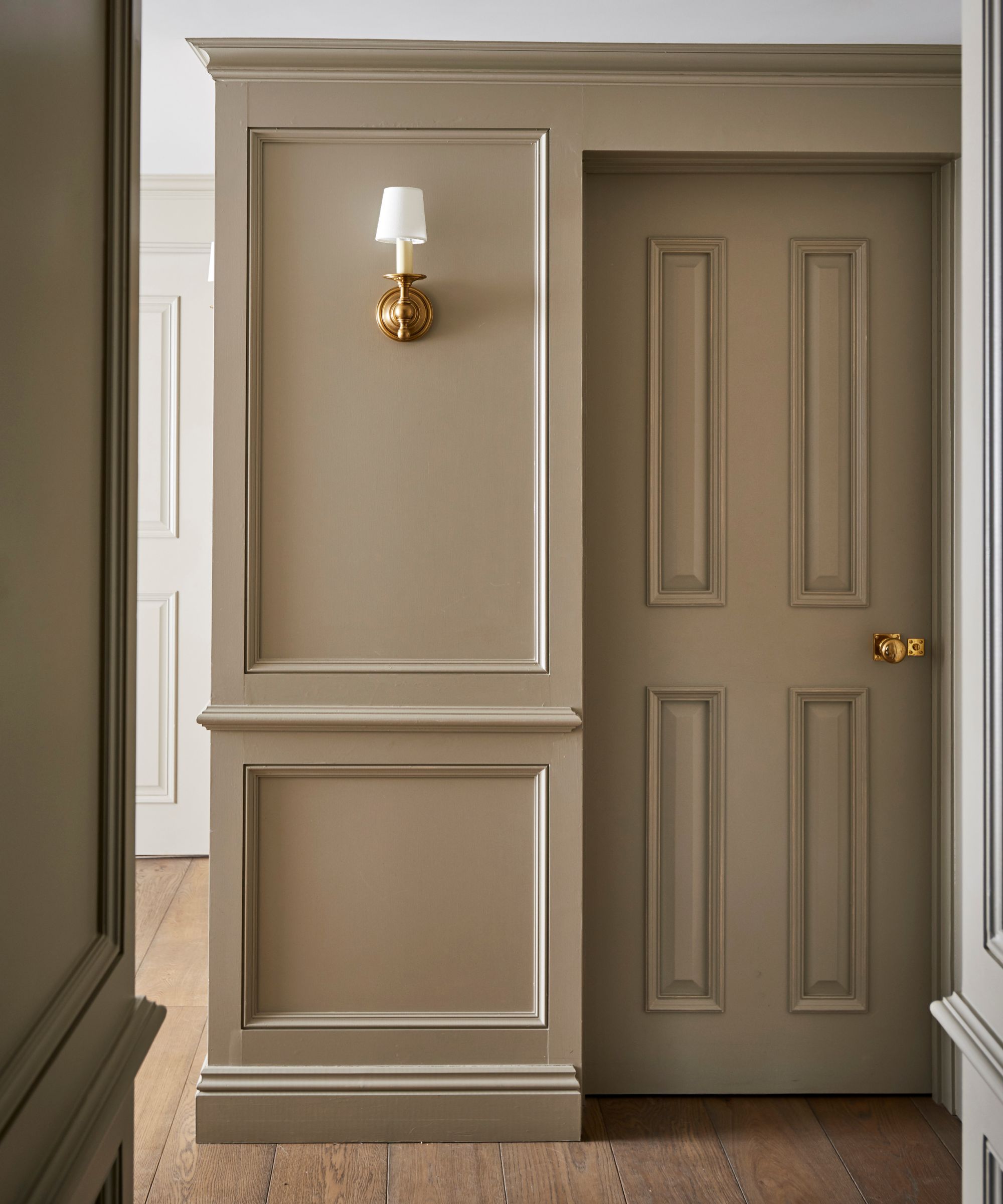 A refined hallway showcasing elegant wall paneling in soft beige tones with intricate detailing. The paneled door blends seamlessly into the design, while a brass wall sconce with a white shade adds a classic touch.