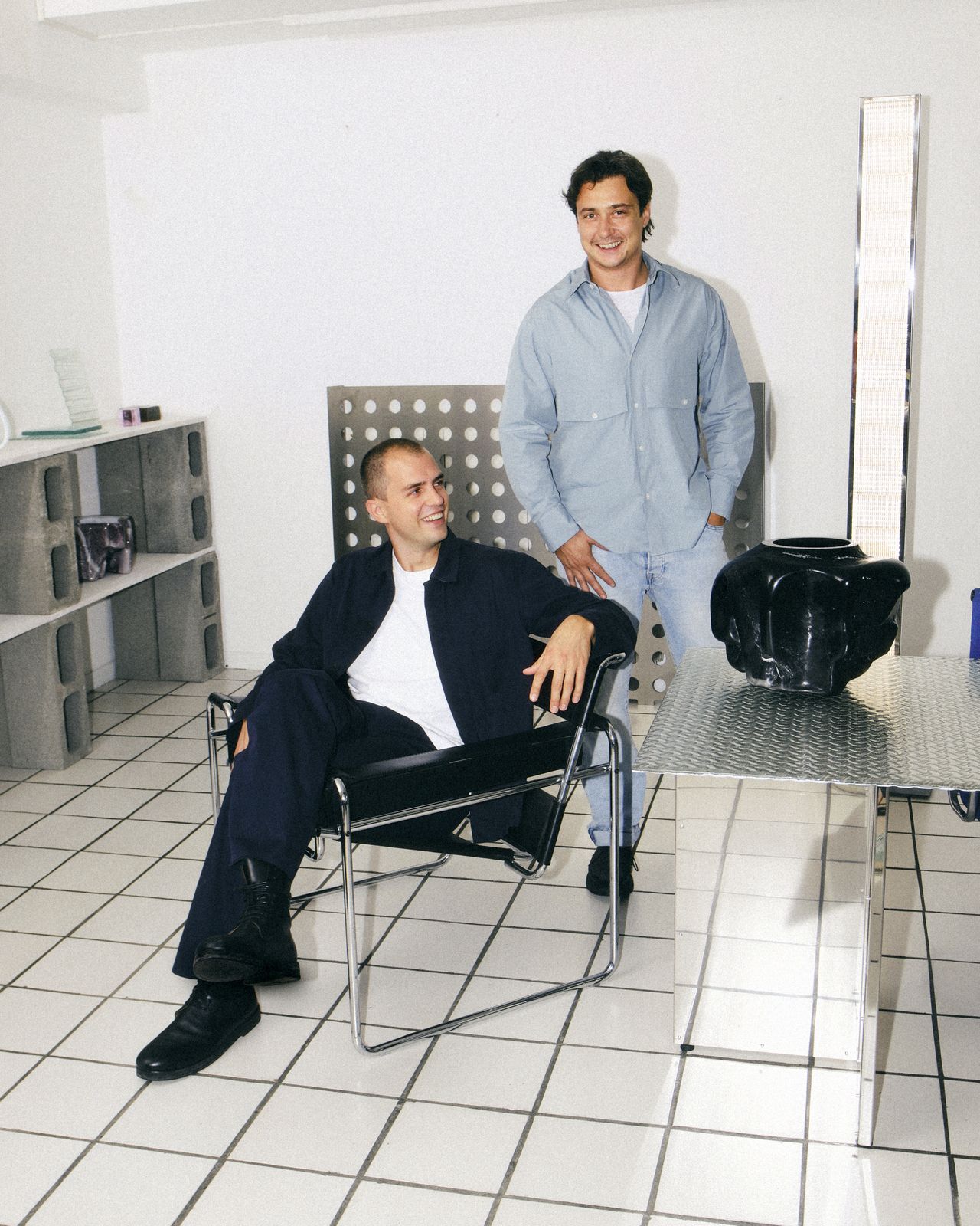 Two young men dressed in all-denim outfits with white T-shirts pose in a modern design showroom filled with chrome items.
