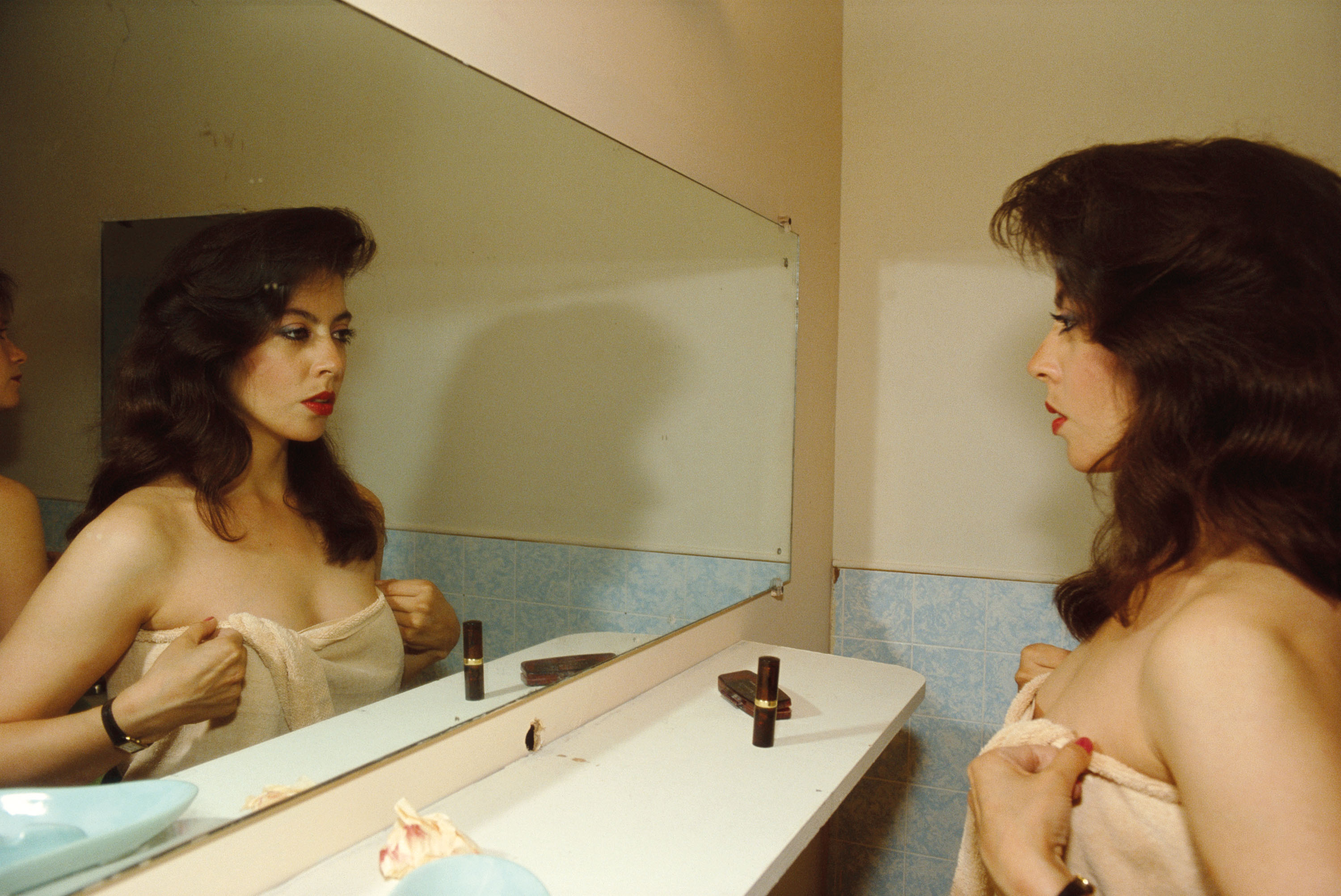 A woman with voluminous dark hair and red lipstick gazes into a bathroom mirror while holding a towel around her body.