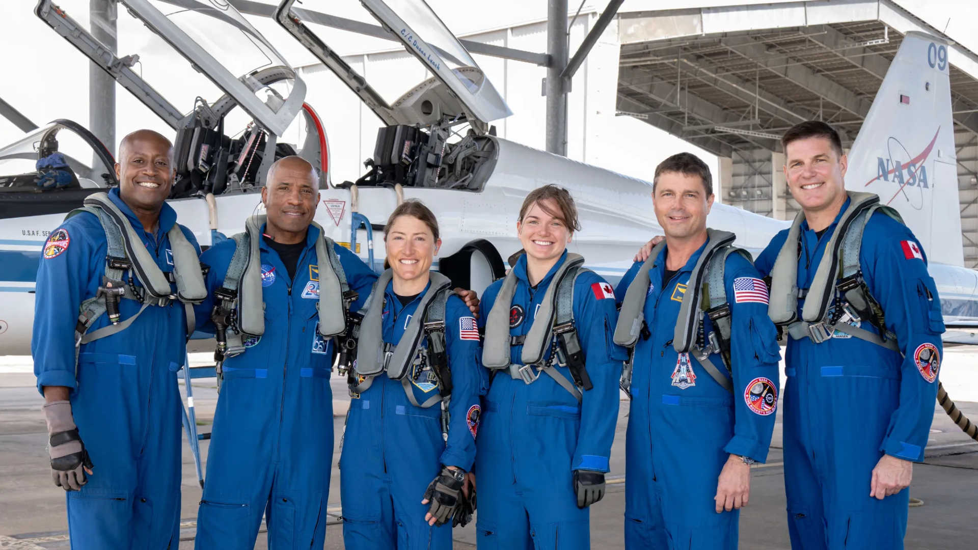 Six people stand in blue jump suits in front of two planes with open cockpits.