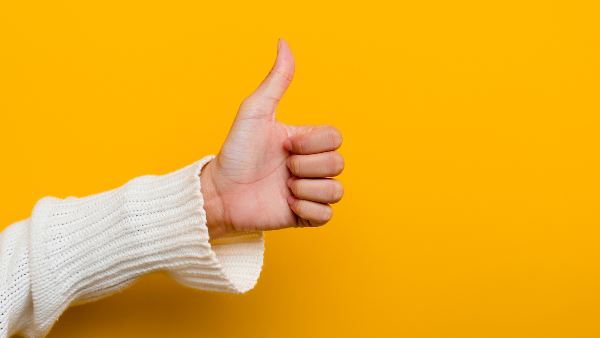 A woman's hand in a thumbs-up gesture against a yellow background.