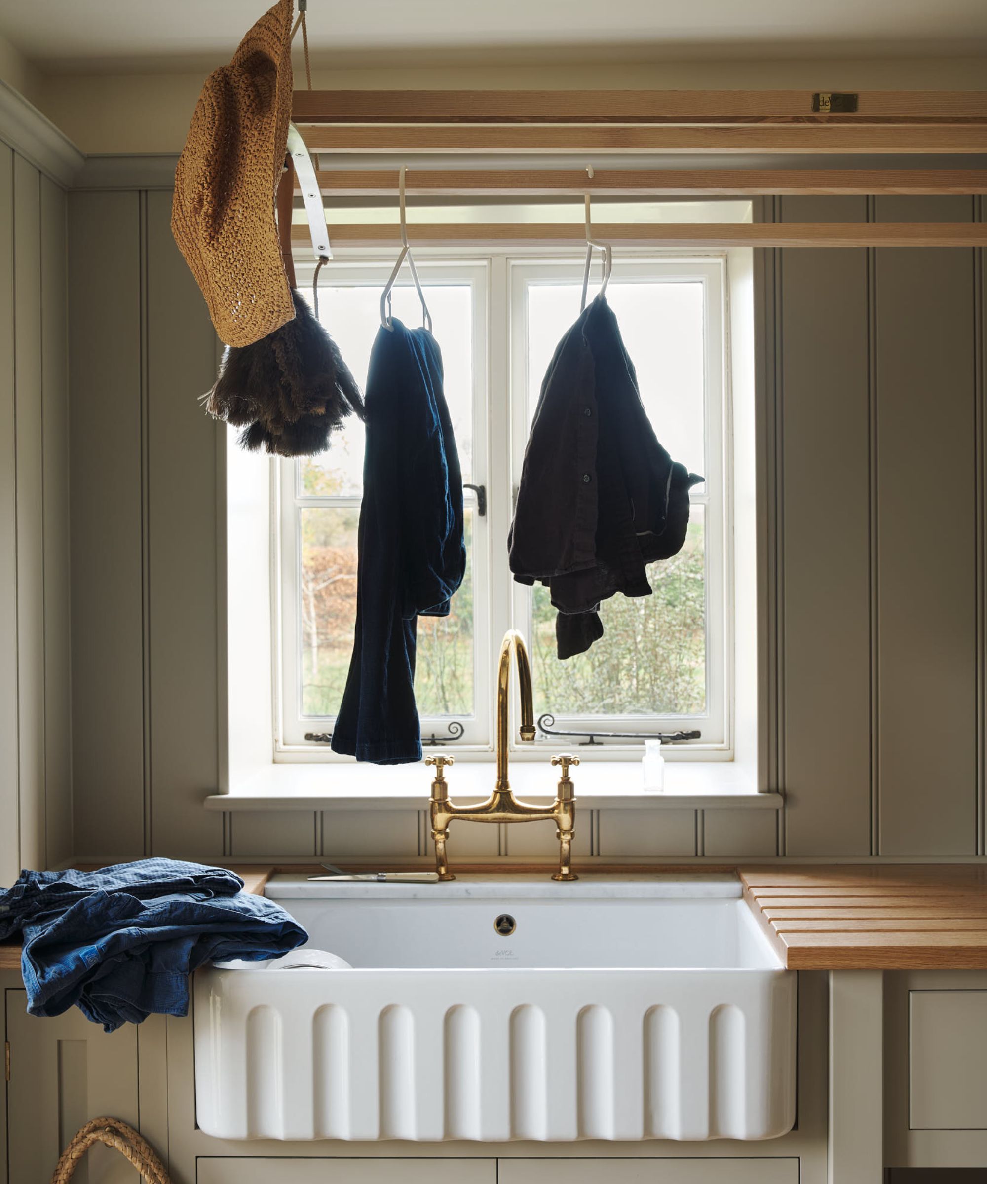 A country utility room with a large Belfast sink, brass bridge tap, oak draining board, ceiling-mounted wooden airer with hanging clothes, and sage green tongue-and-groove panelling.