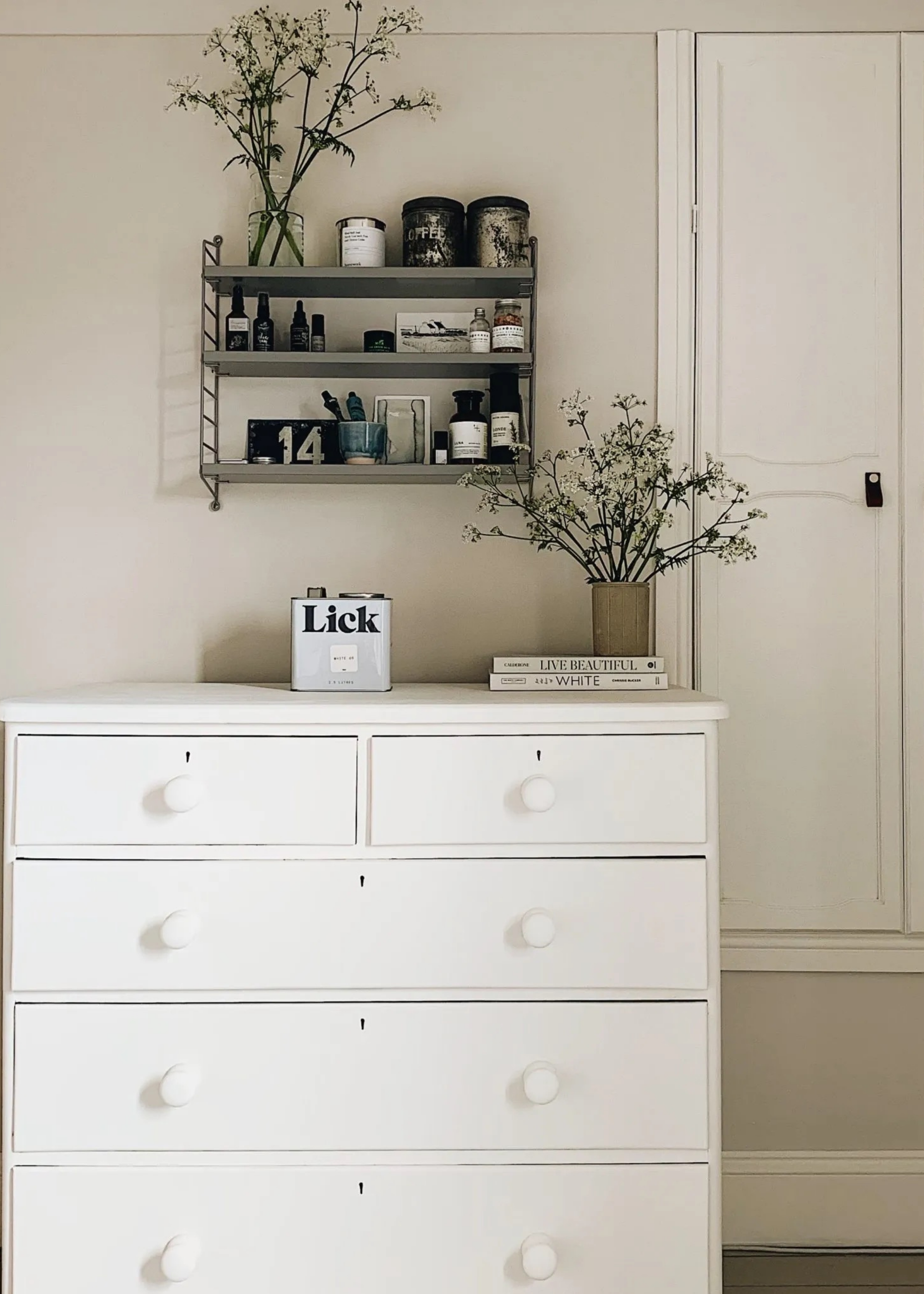 room with off-white walls, white painted cabinet, with floating shelves styled with trinkets, a can of lick paint on the dresser, and two books topped with a vase of flowers
