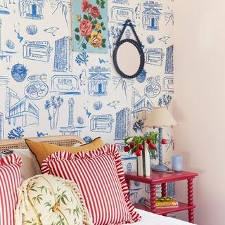 A bedroom with a wall covered in a hand-drawn wallpaper with a red bobbin bedside table and striped, frilled cushions on the bed