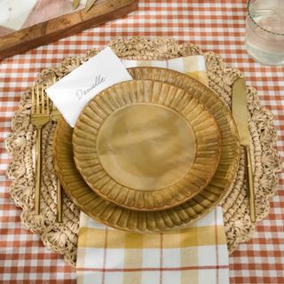 A yellow magnolia dinner plate with a smaller serving plate on top, gold cutlery on either side, a woven placemat, and red and white gingham tablecloth underneath