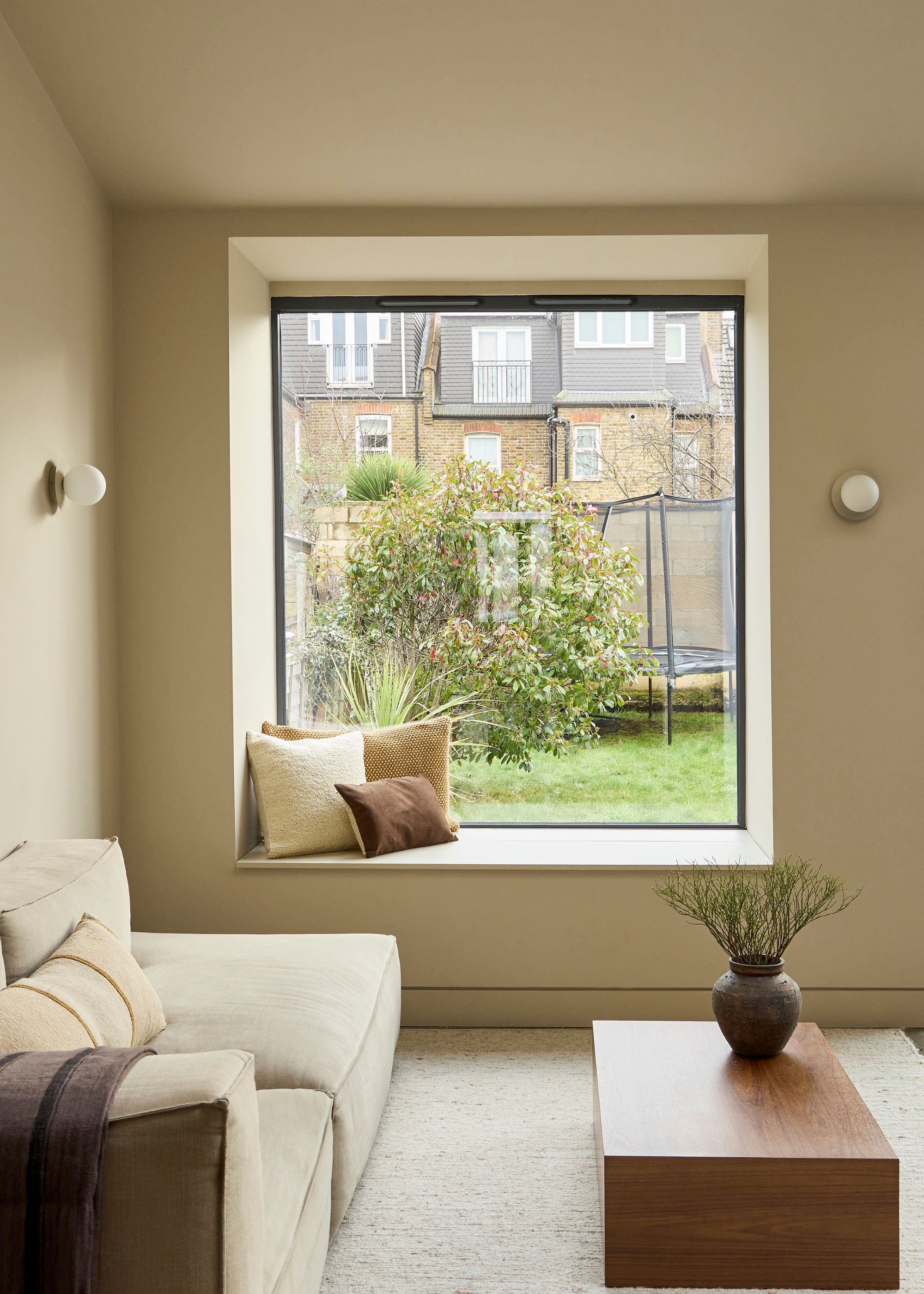 A modern extension with a neutral-hued seating area with a beige sofa and low wood coffee table and a window seat shows a view of the garden with a trampoline