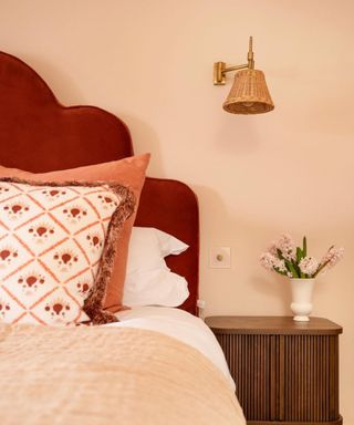 bedroom with light pink wall, red wavy headboard and soft pink bedding with wooden nightstand and woven wall sconce