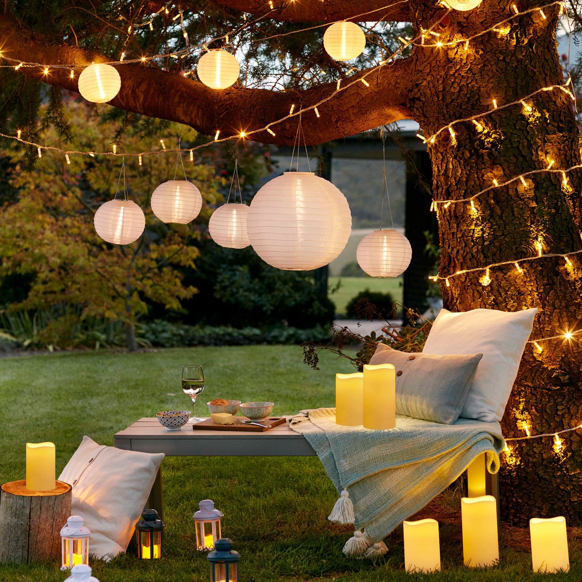 A garden with a tree decorated with fairy light hung on a tree and lantern lights suspended from a large branch hovering above a sun lounger