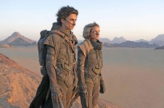 timothee chalamet as paul atreides and rebecca ferguson as his mom wearing space suits and standing on a sand dune in the movie dune