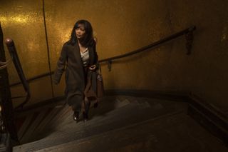 Celia (Thandiwe Newton) is ascending a spiral staircase with bannisters on both sides. The tiles on the walls behind her are bronze and gold-coloured. She is wearing a long coat, carrying a handbag in the crook of her left arm, and looks apprehensive