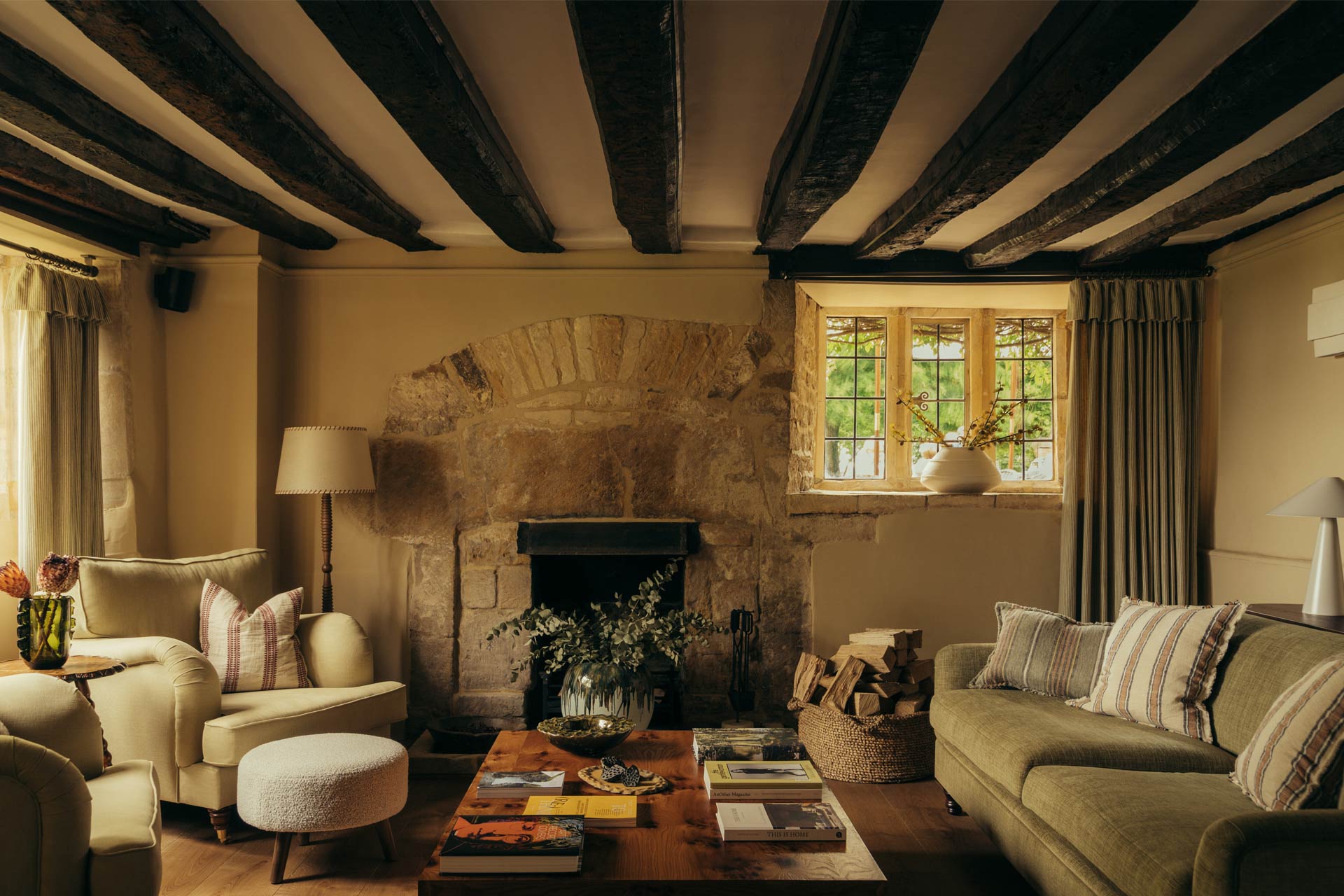 A vibrantly decorated farmhouse with plush fabrics, exposed wooden beams, a stone-clad fireplace, colorful books, and creamy furniture.