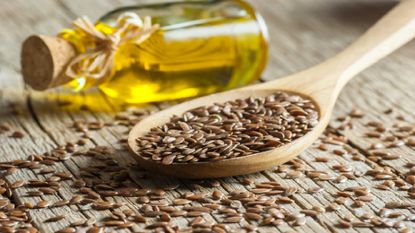 Flaxseed in a wooden spoon next to a small glass bottle on its side filled with oil
