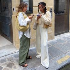 fashion influencer Amaka Hamelijnck and a friend poses on the side walk of a city street in stylish casual neutral outfits while looking at their cell phones.