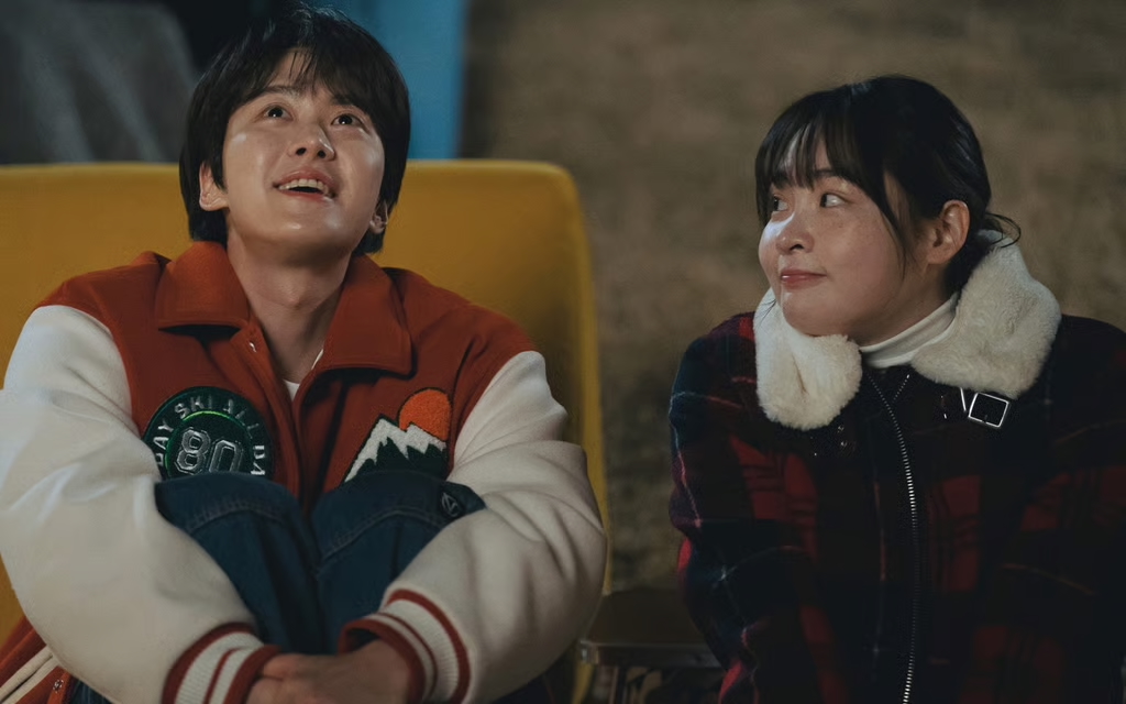 A Korean man in a letterman's jacket (Gong Myung) looks at the sky while sitting in a yellow slide, as a Korean woman in a fur-lined jacket (Kim Min-ha) sits to his right and looks at him. A still from the K-drama 'Way Back Love.'