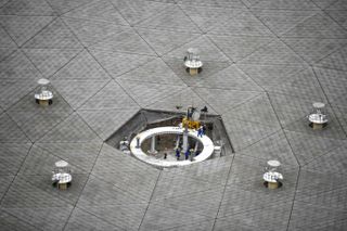 In the bottom of an enormous radio telescope dish, small workers are seen like ants, performing maintenance.