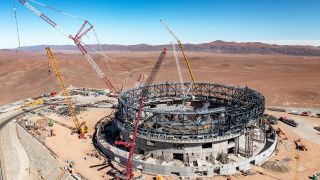 A large steel frame is being put together by large yellow and red cranes.