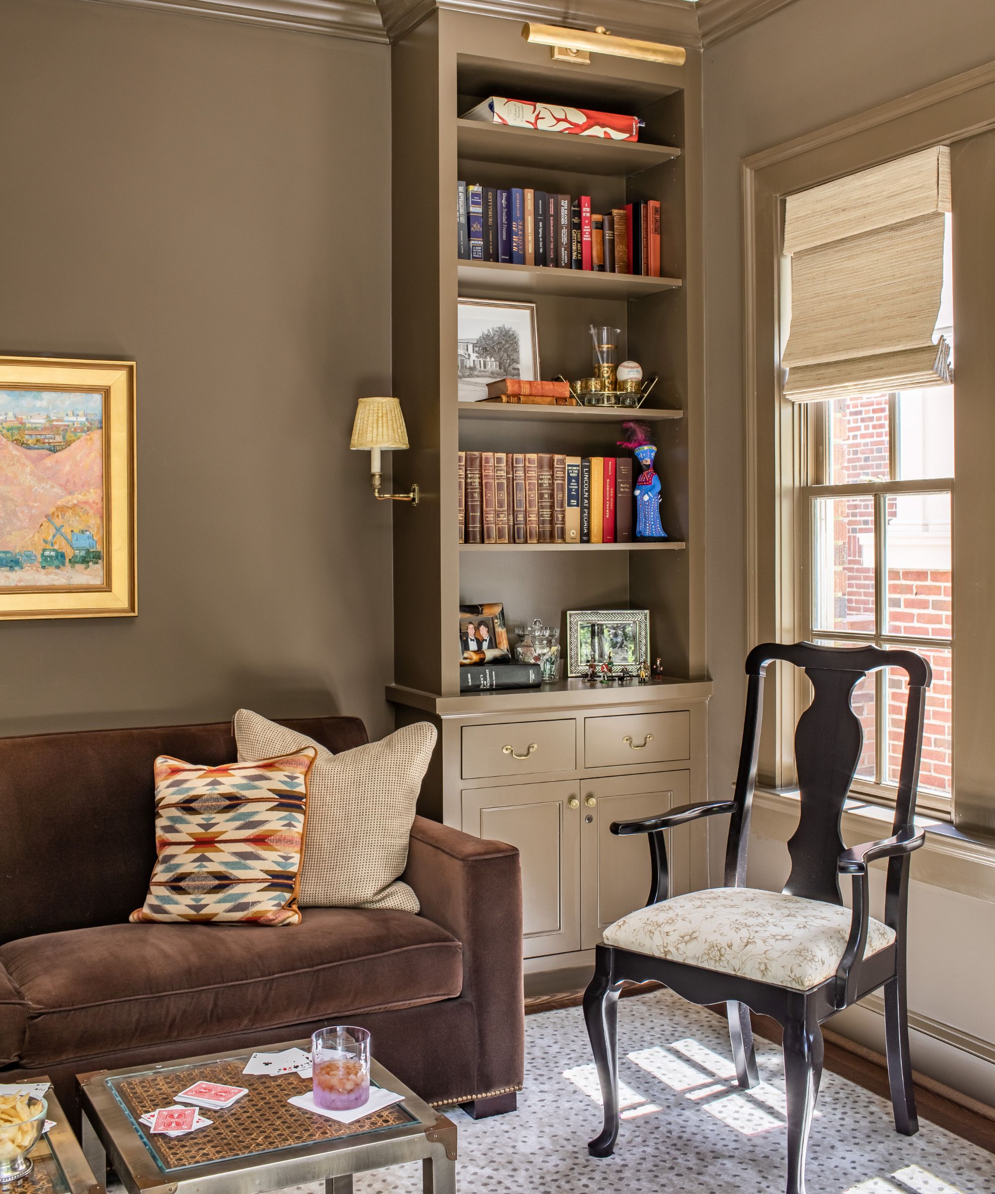 Sitting room with brown painted walls and a bookcase, a brown velvet sofa, a rattan coffee table, a vintage chair, a polka dot rug and a vintage painting
